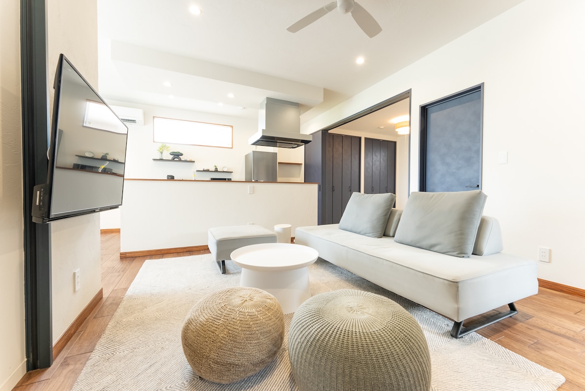 A modern living area is highlighted by a comfortable sofa and casual poufs. A wall-mounted television faces the seating area, while natural light illuminates the room through a large window. The kitchen appears in the background, creating an inviting, open layout.