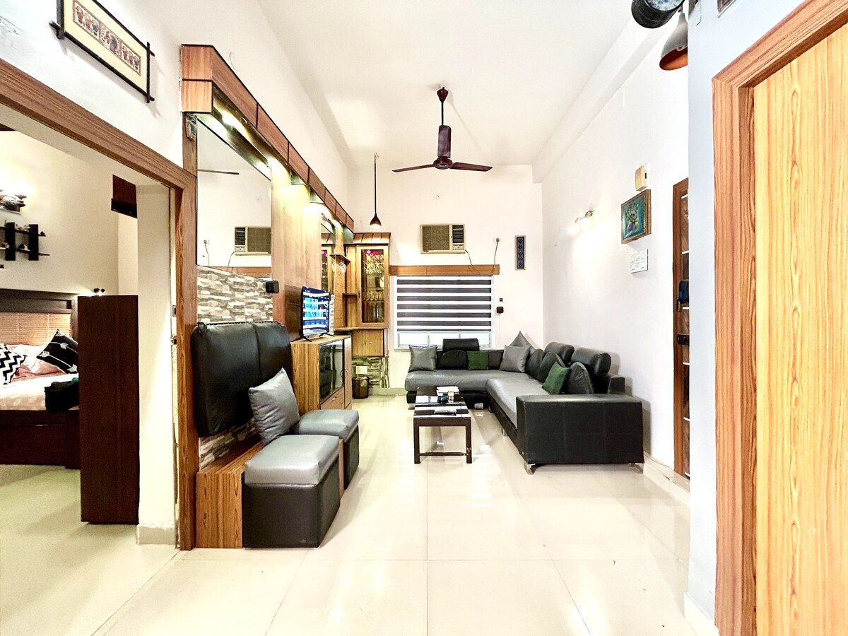 A spacious living area features a comfortable arrangement of a sectional sofa and wooden furnishings. Natural light fills the room, highlighting the sleek tiled floor. An artistic display is mounted on the wall, complementing the modern decor, while a ceiling fan provides ventilation.