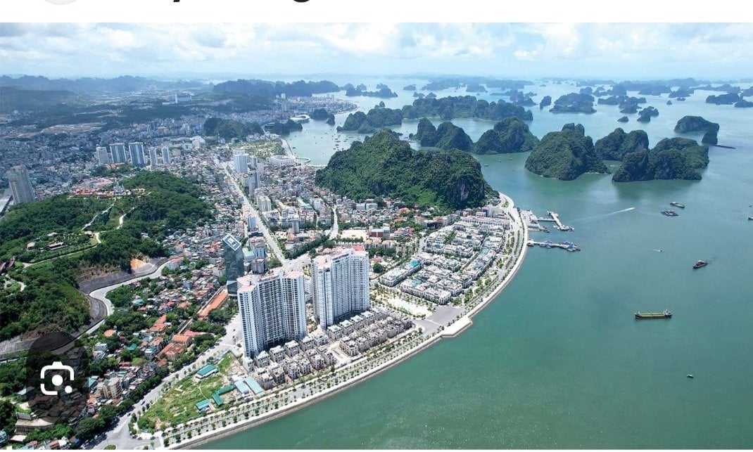 An aerial view captures the layout of a coastal city set against lush green mountains and the tranquil waters of Ha Long Bay. Tall buildings line the waterfront, while smaller structures are surrounded by greenery, showcasing a blend of urban and natural environments.