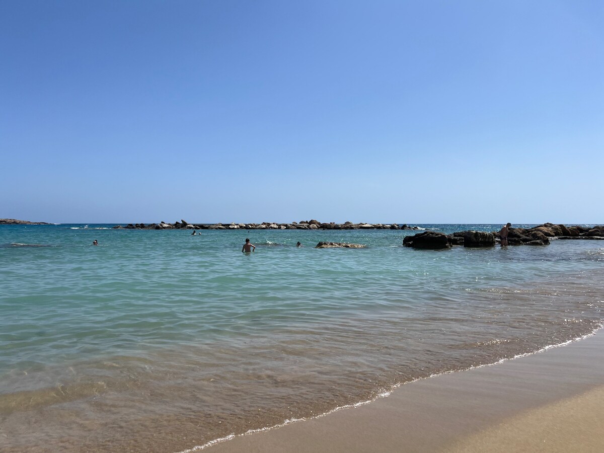A serene beach scene features calm turquoise waters gently lapping against a sandy shore. In the distance, swimmers can be seen enjoying the clear water, while rocky formations outline the horizon under a clear blue sky.