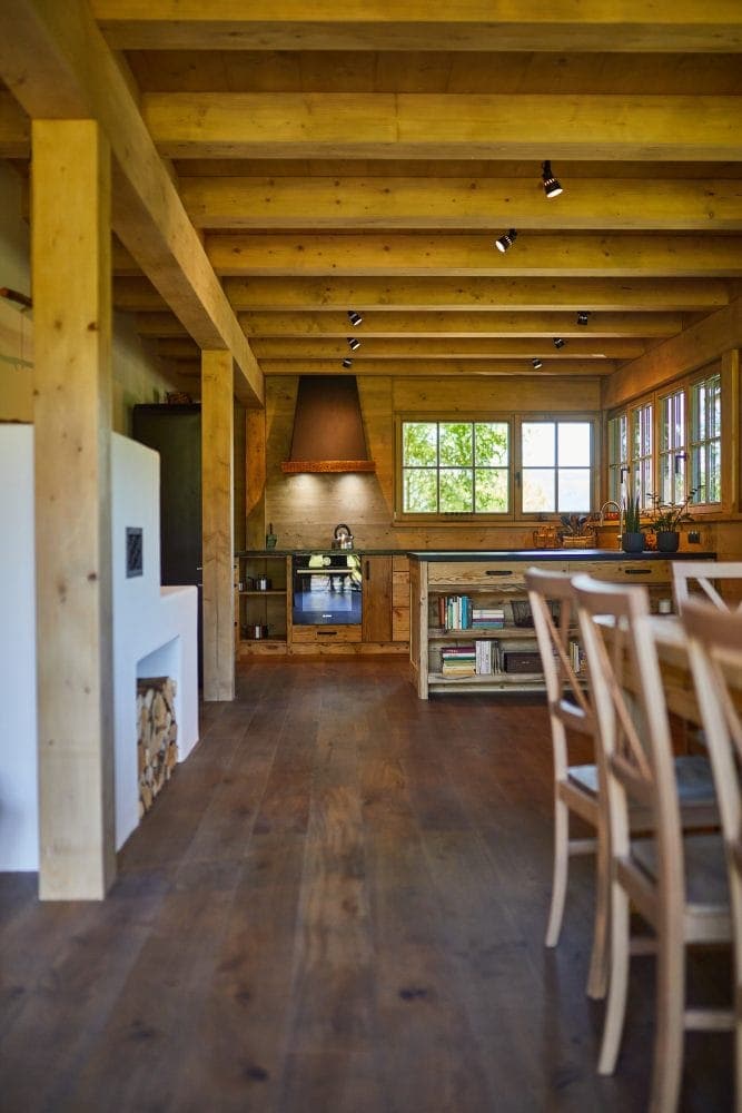 A warm kitchen area features wooden beams overhead and a rustic design. Light streams through multiple windows, illuminating the wooden cabinetry and countertops. A fireplace is visible to one side, with neatly stacked firewood nearby. A dining table with several chairs is positioned towards the foreground.