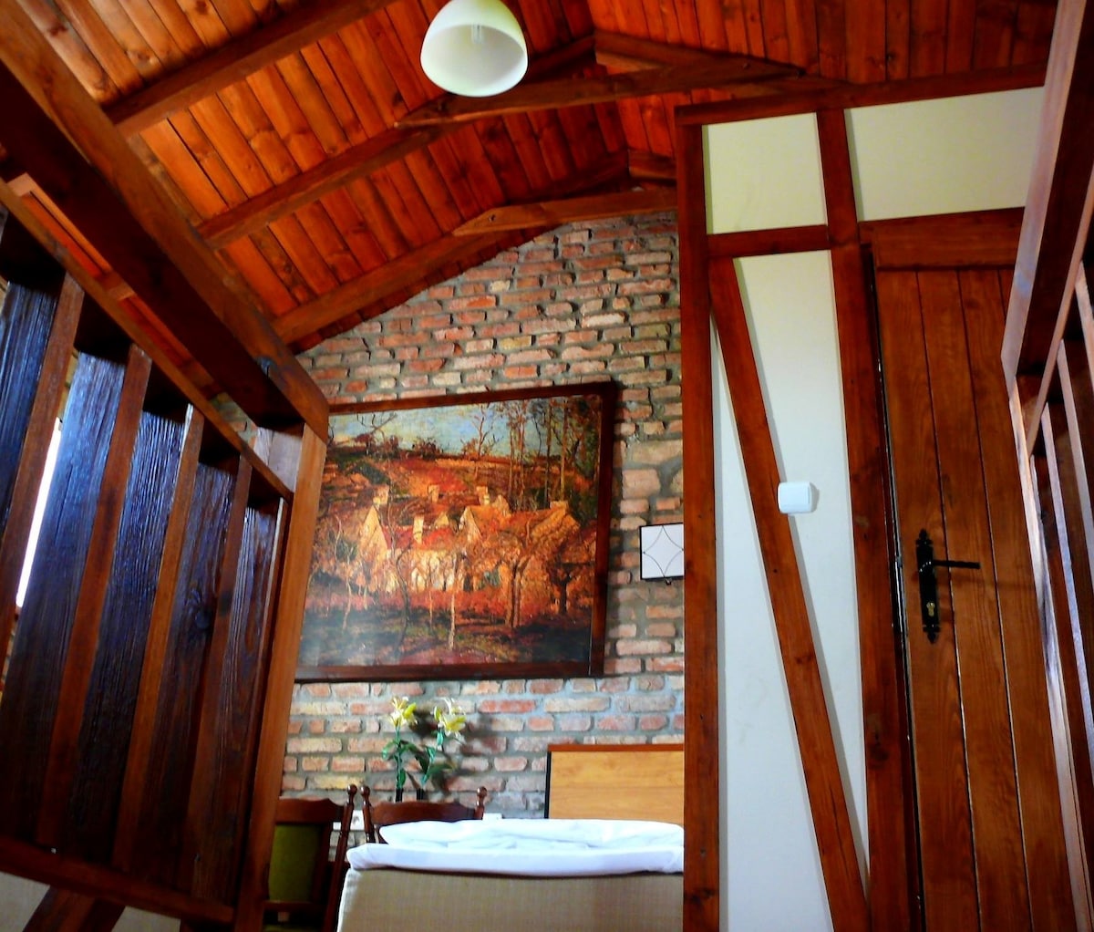 A cozy interior space features exposed wooden beams and a brick wall, complemented by a large artwork depicting a rural scene. Natural light enters through a small window, illuminating the simple furnishings, including a bed with a neutral-colored bedding.