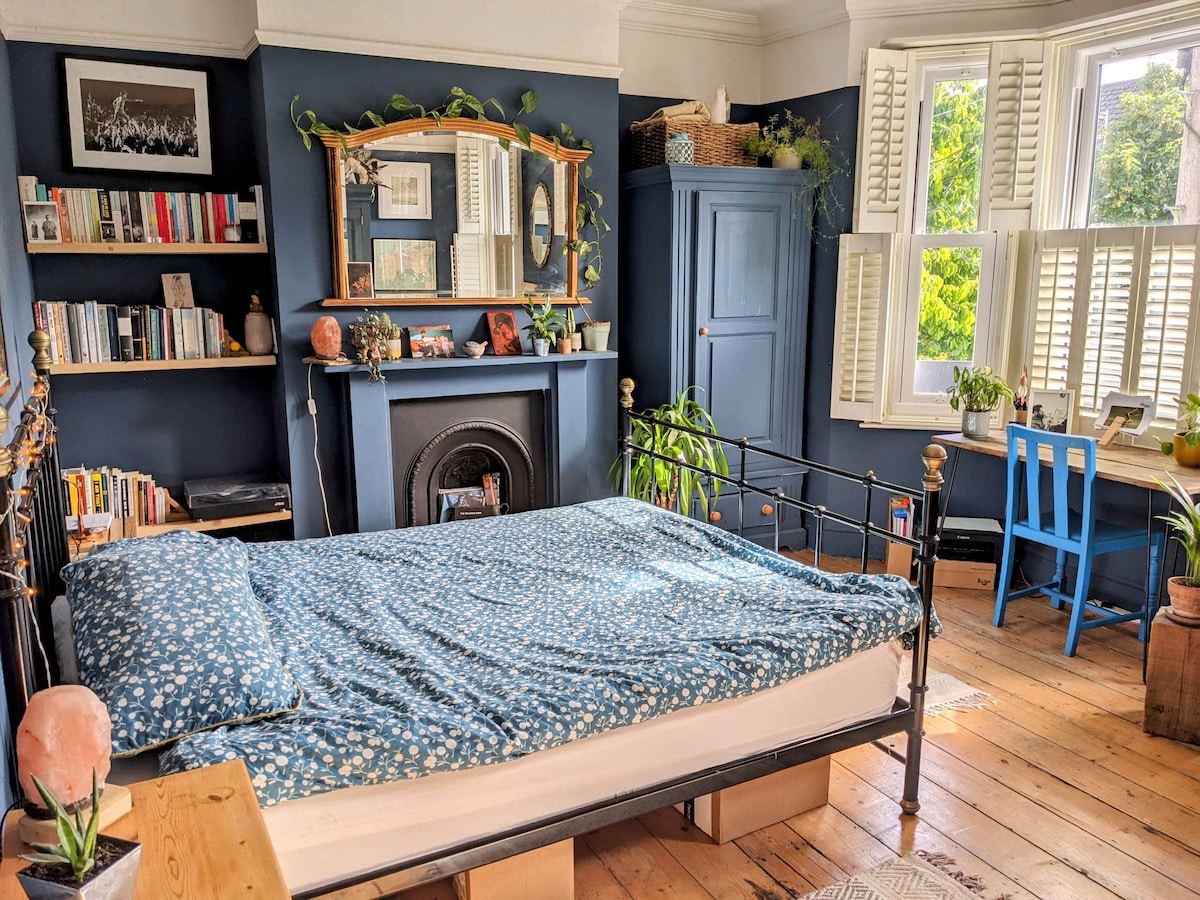 A spacious bedroom features a king-sized bed covered with a floral blue blanket. A bookshelf and a desk are present, along with decorative elements on the mantelpiece. Bright light enters through the large window, complemented by elegant shutters.