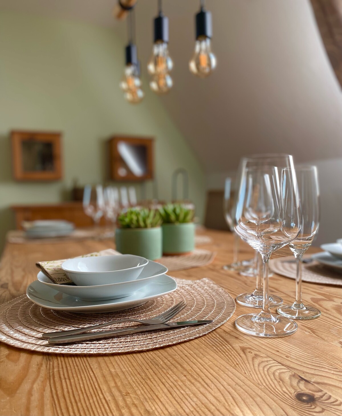 A dining table is set with neatly arranged plates, cutlery, and glasses. Soft green walls provide a backdrop for the decor. A small plant is placed in the center. A warm natural light filters in, enhancing the inviting table setting.