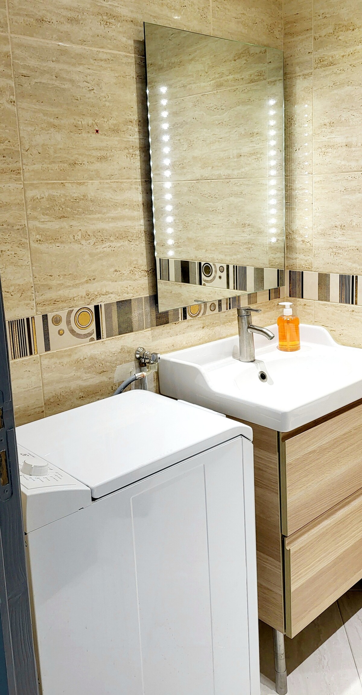 A modern bathroom includes a sleek vanity with a white sink positioned next to a washing machine. A large mirror reflects warm lighting, while a decorative tile accent runs along the wall. Storage drawers are provided beneath the sink for convenience.