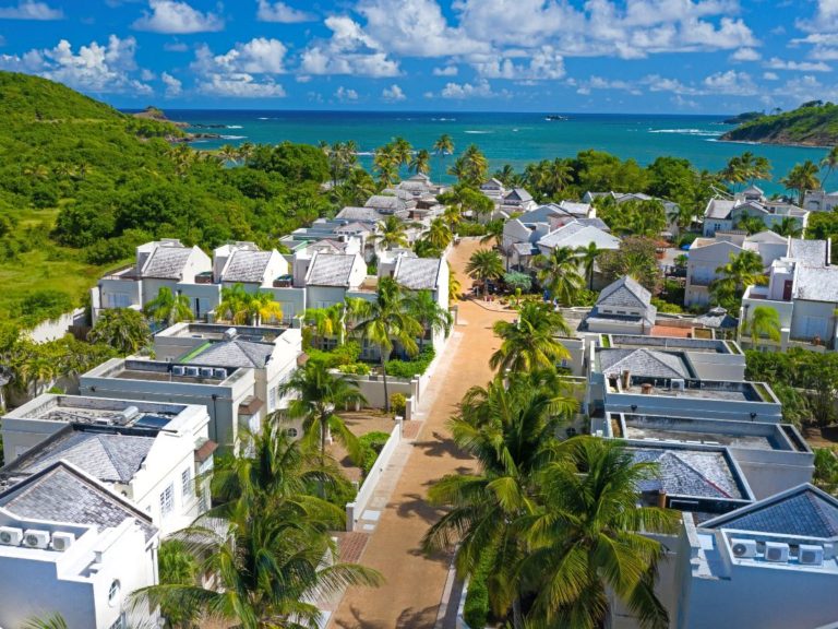 An aerial view captures a peaceful community surrounded by lush greenery, leading towards the ocean. White rooftops dot the landscape, with palm trees lining the pathway. The clear blue waters and tropical scenery create a serene backdrop for this coastal residential area.