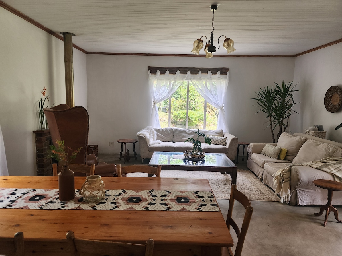 A spacious living area is featured, showcasing a wooden dining table with several chairs and a decorative table runner. A comfortable seating arrangement includes a light-colored sofa and a vintage armchair. Natural light is provided by a large window adorned with sheer curtains.