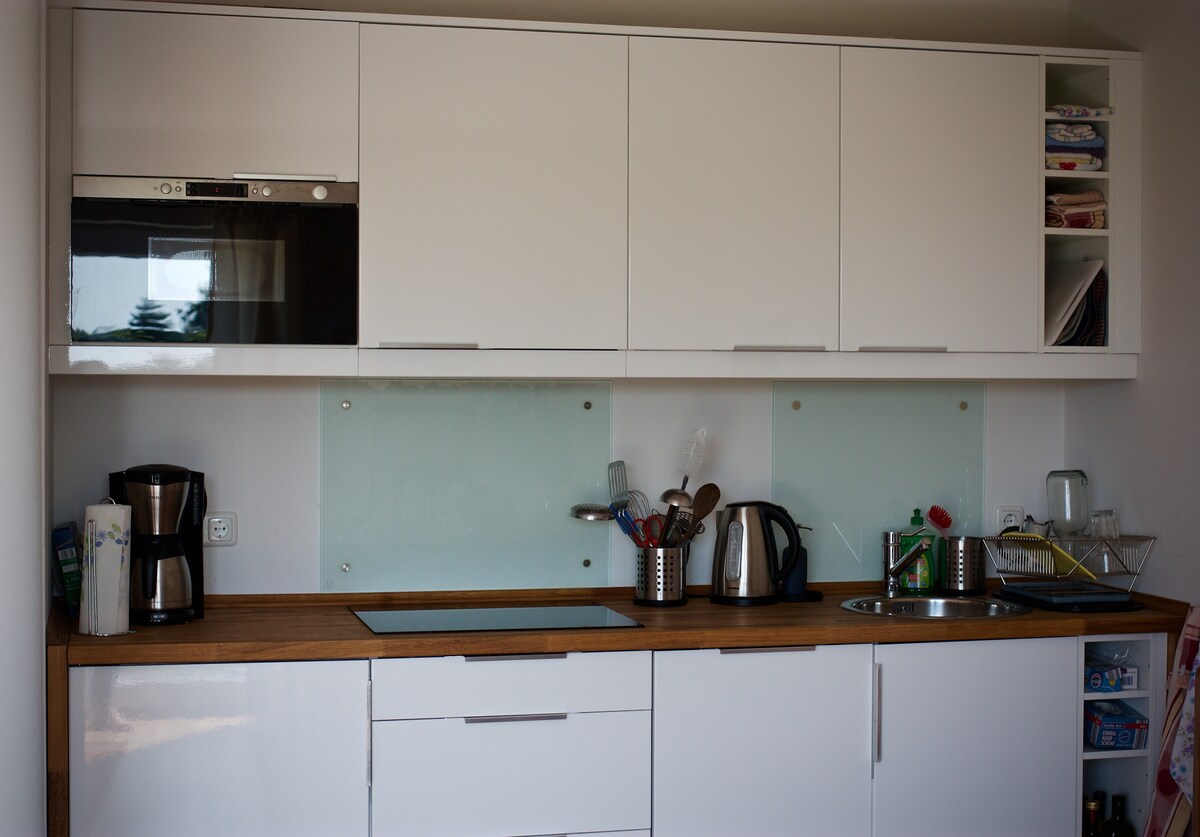 A modern kitchen is displayed with sleek white cabinetry and a wooden countertop. An integrated microwave is positioned above the cooking area. Various kitchen appliances, including a coffee maker, kettle, and utensils, are organized neatly on the countertop, emphasizing functionality and ease of use.