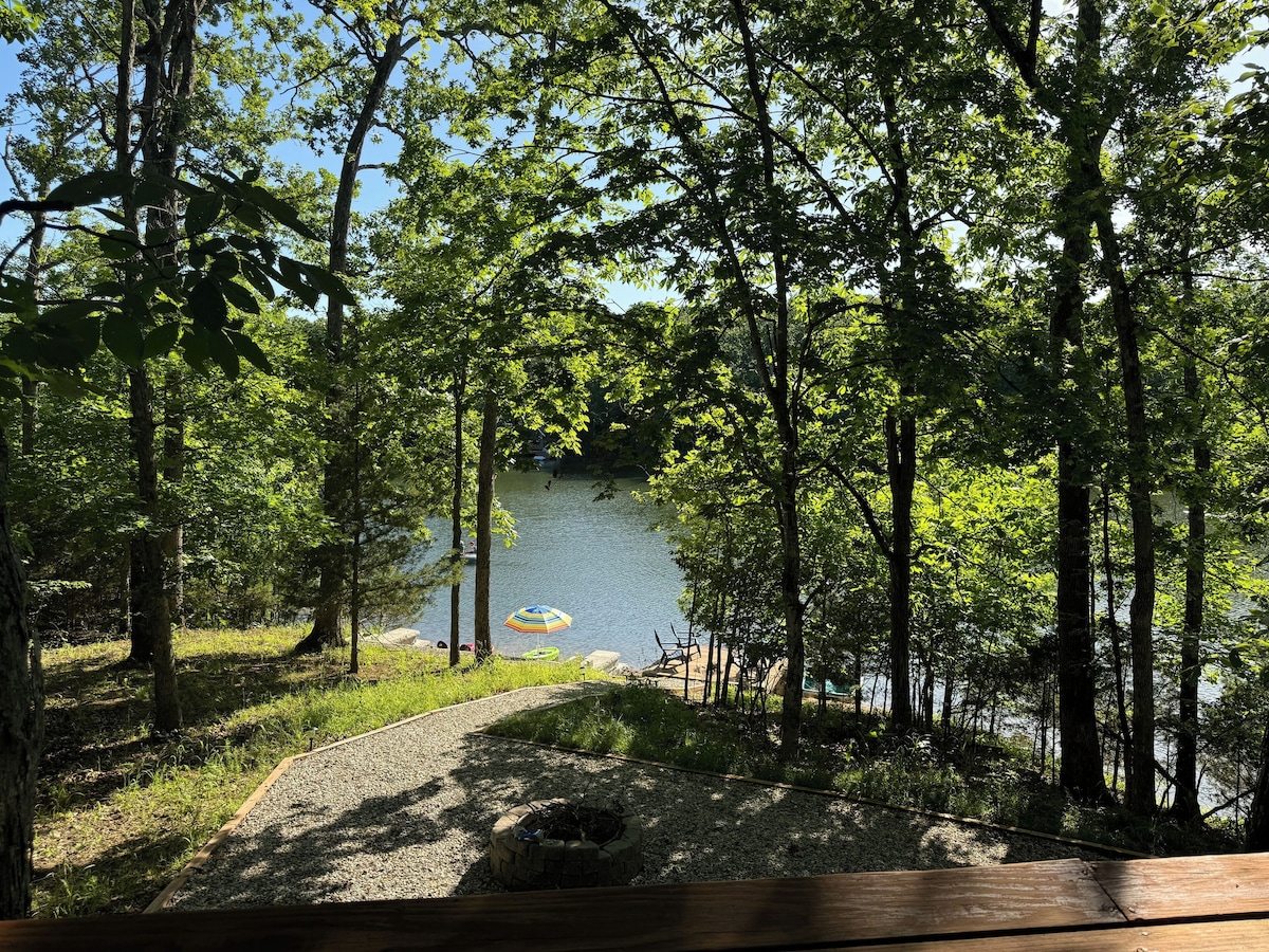 A serene view of the lake is framed by lush green trees, with a private beach visible nearby. A circular fire pit is set on gravel, and a paddle boat floats gently on the water, creating a calm atmosphere.