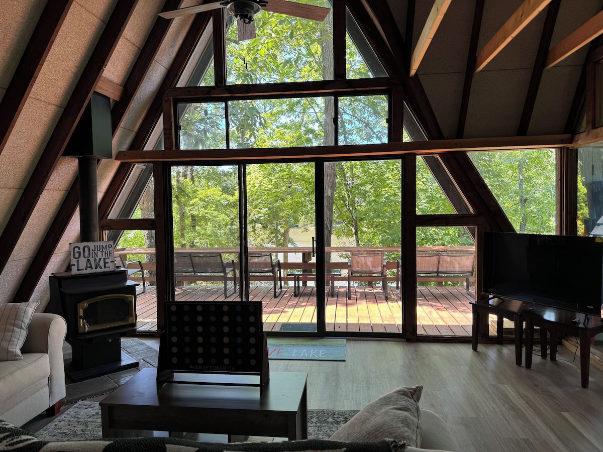Large windows allow natural light to fill the living area, providing views of the surrounding trees. A comfortable seating arrangement includes a couch and armchair, while a wood stove adds a cozy touch. A television sits in the corner, completing the relaxing interior space.