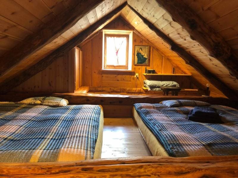 An attic space features two spacious beds dressed in checkered bedspreads. The walls are lined with warm wood paneling, while a small window allows soft light to enter. A framed artwork and a small shelf are visible, adding subtle character to the room.