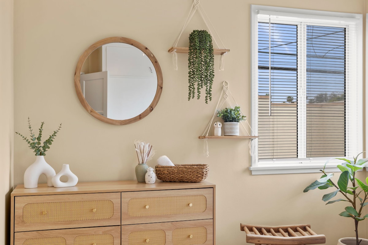 A light-colored room features a wooden dresser with woven detailing, accompanied by a round mirror and decorative shelves. Small potted plants and a vase with greenery are displayed on the shelves, while a window with white blinds allows natural light to brighten the space.
