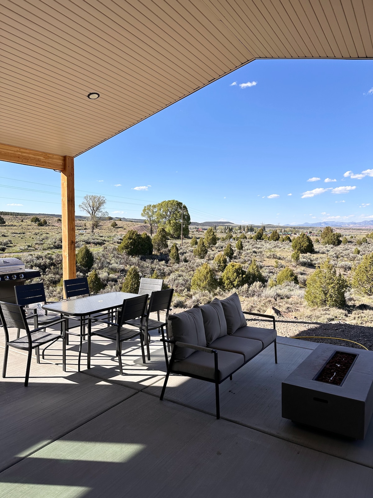 An outdoor patio area is showcased, featuring a dining table with seating for six, accompanied by a comfortable sofa. A barbecue grill is positioned nearby, and the scenic landscape reveals shrubs and open spaces under a clear blue sky.