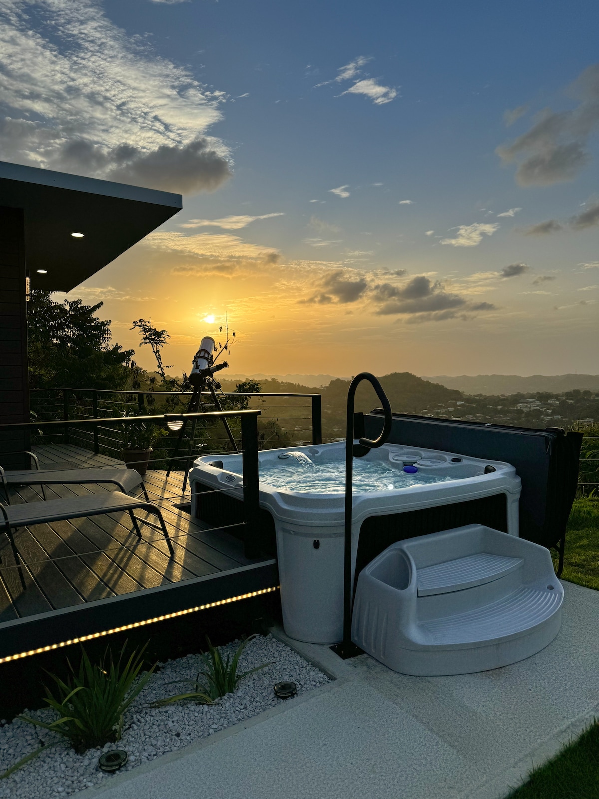 A hot tub is positioned on a wooden deck, overlooking expansive hillside views during sunset. The scene is complemented by a telescope ready for stargazing. Soft lighting highlights the edges of the deck, enhancing the tranquil outdoor setting.