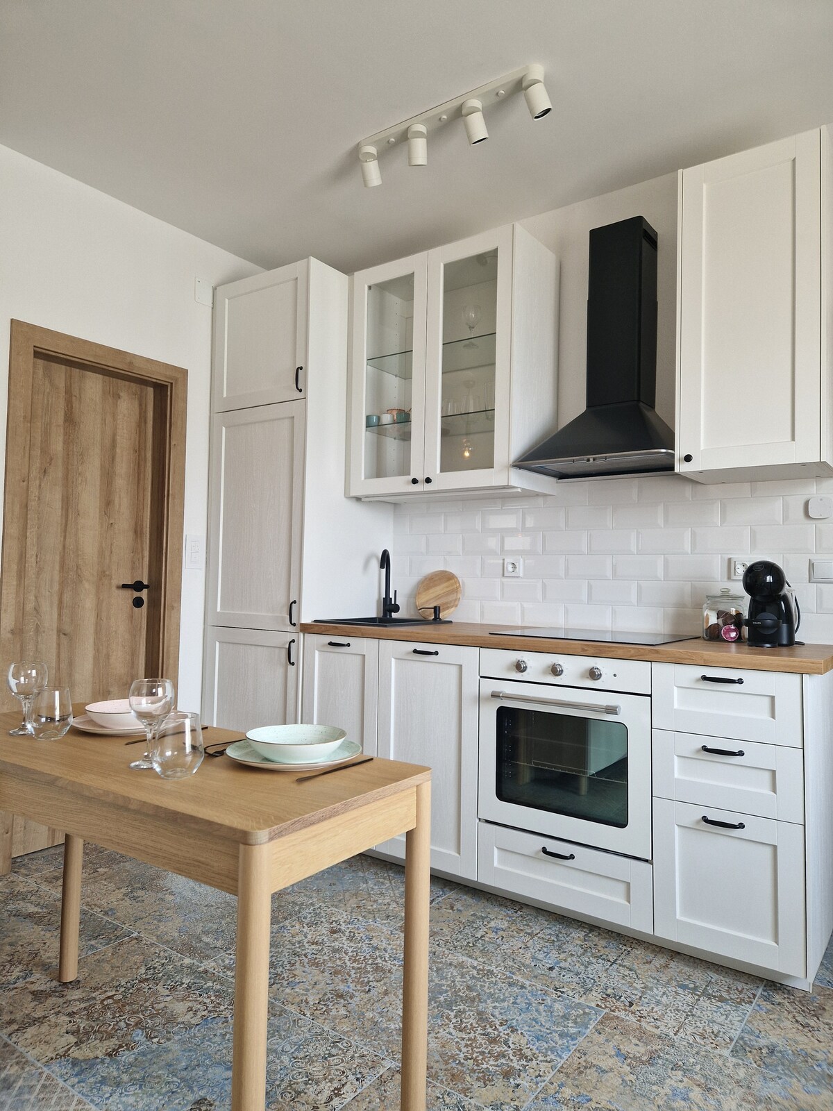 A modern kitchen features sleek white cabinetry and a wooden countertop. An oven and a minimalist sink are integrated into the cabinetry. A small dining table is set with plates and glasses, creating an inviting space for meals. Soft light is provided by track lighting above.
