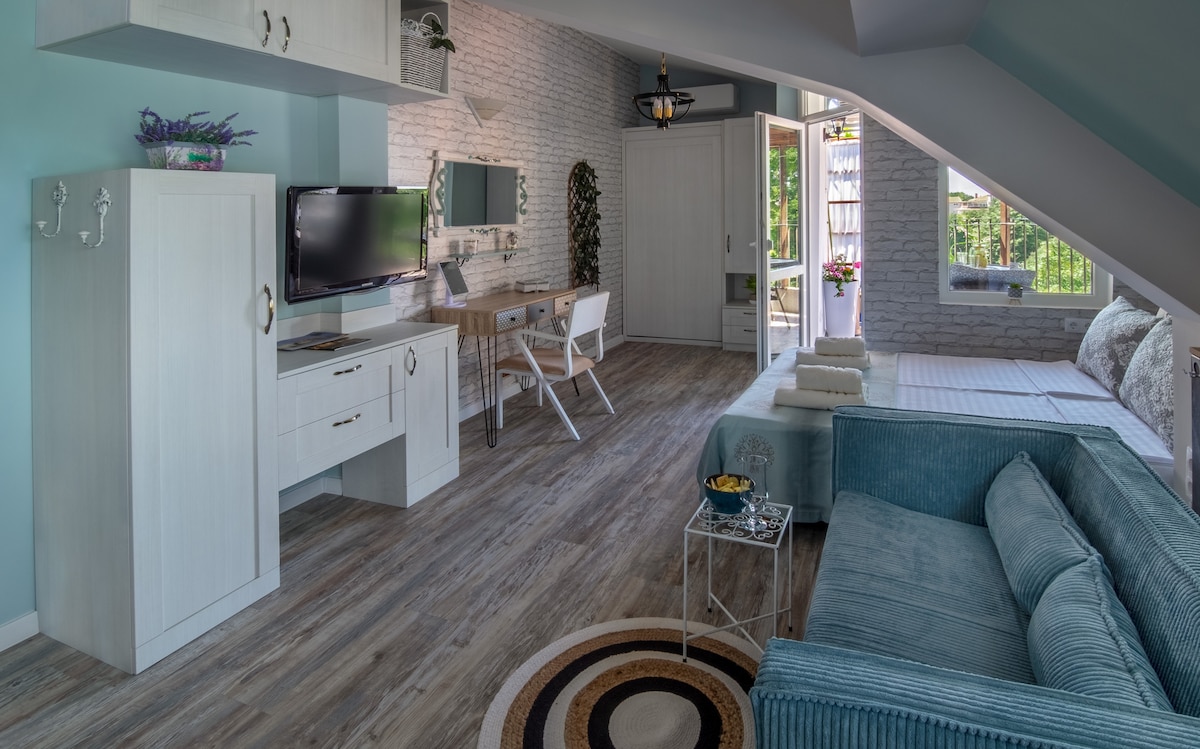 A bright studio features custom-made furnishings, including a cozy sofa and a desk. A television is mounted on the wall. The space is complemented by wooden flooring and elegant light fixtures, with a terrace visible through the open glass doors.