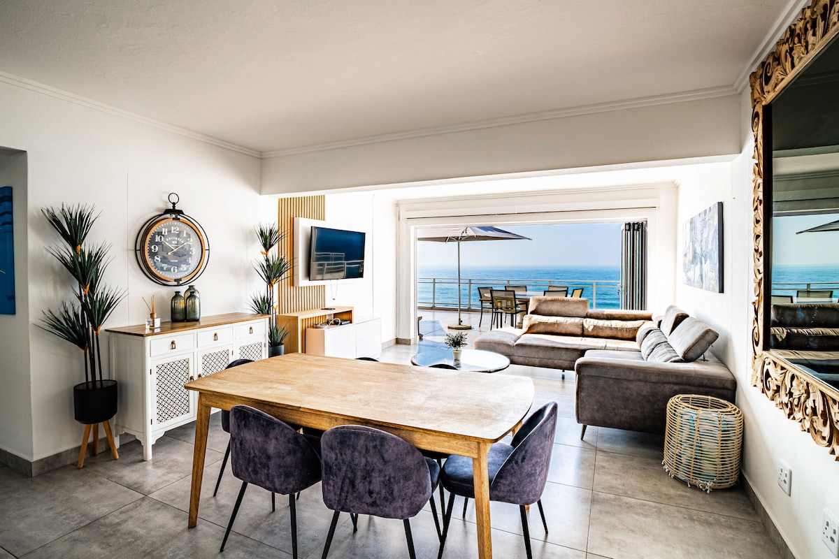 An airy living area showcases a wooden dining table surrounded by upholstered chairs. A modern sofa and side table are positioned near expansive glass doors that open to reveal views of the ocean. Decorative plants and a wall mirror enhance the elegant atmosphere.