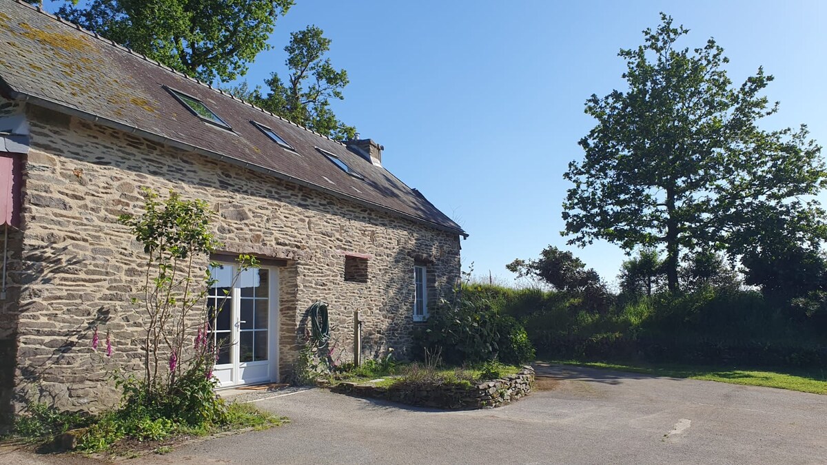 An attractive stone building is featured, showcasing a rustic facade with a sloped roof. The entrance is framed by plants and a small garden area. A wide driveway leads to the entrance, while green trees are visible in the background under a clear blue sky.