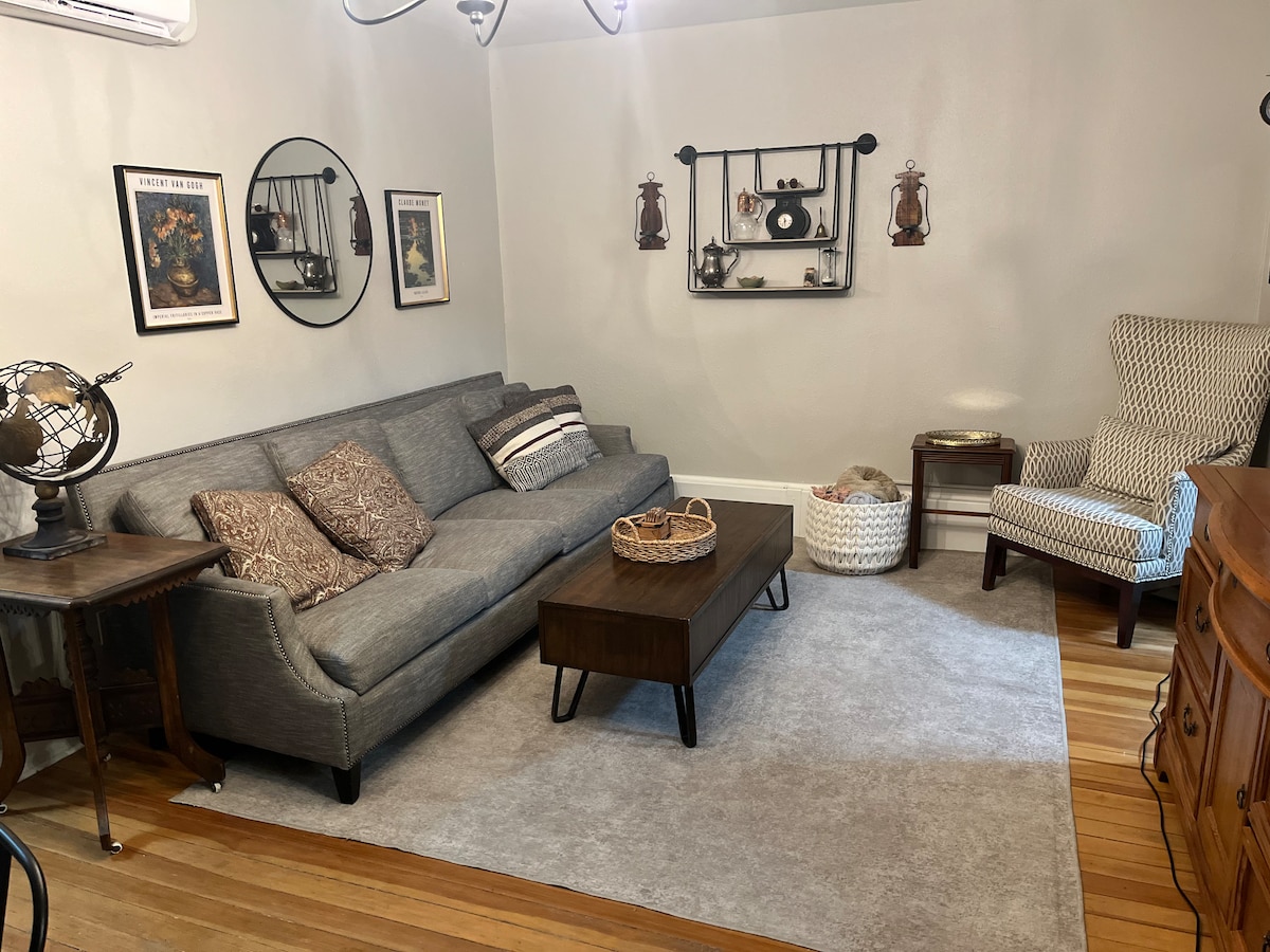 A cozy living room features a gray sectional sofa adorned with decorative pillows. A wooden coffee table sits at its center, accompanied by a decorative bowl. On the walls, vintage artwork and shelves with lanterns add character. A patterned armchair offers additional seating beside a textured area rug.
