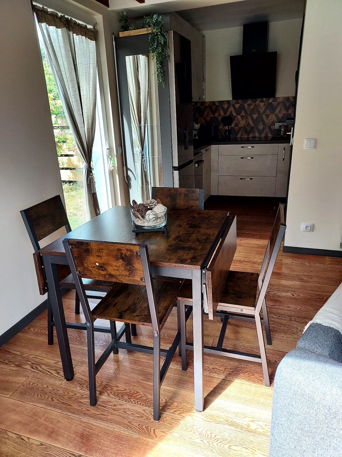 A modern dining area features a wooden table accompanied by four matching chairs. Natural light enters through glass doors that open to the outdoors, enhancing the spacious feel. A decorative centerpiece rests on the table, adding a touch of detail to the functional space.
