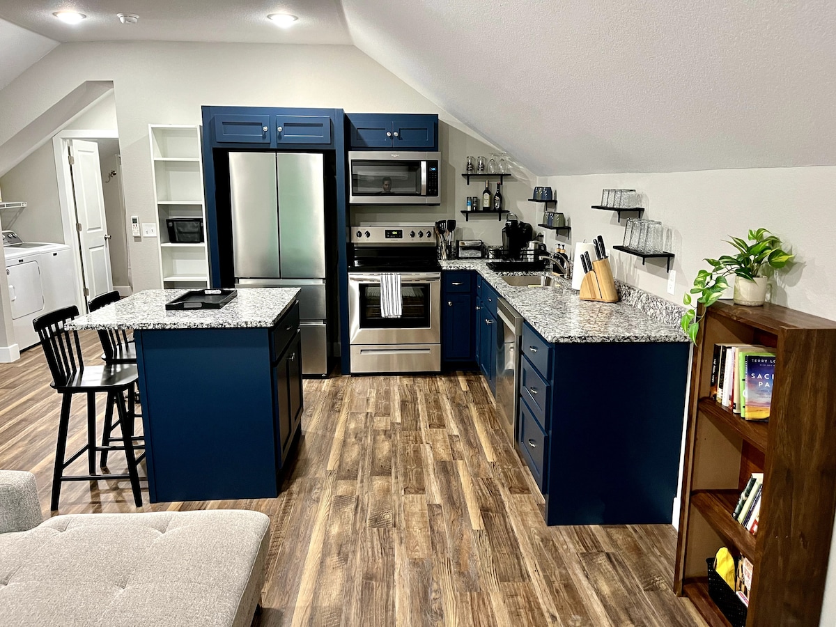 The kitchen features modern blue cabinetry and granite countertops, equipped with stainless steel appliances, including a stove and refrigerator. An island with seating for two, glassware on open shelves, and a small bookshelf with various titles enhance the functional space.