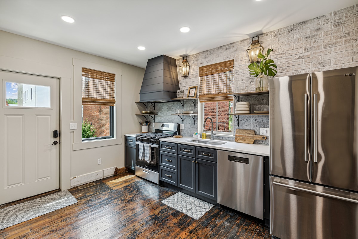 The kitchen features dark cabinetry complemented by a stainless steel refrigerator and oven. A farmhouse sink is positioned beneath a window, letting in natural light. Wood flooring and woven shades enhance the space's warmth, while decorative shelves display curated kitchen essentials.