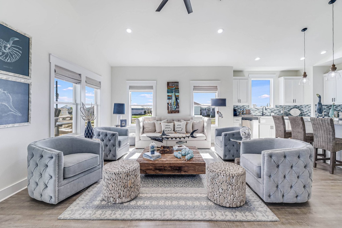 A spacious living area features a light color palette with two elegant armchairs and a comfortable sofa, arranged around a wooden coffee table. Natural light pours in through large windows, showcasing views of the outdoors. Soft decorative accents enhance the inviting atmosphere.