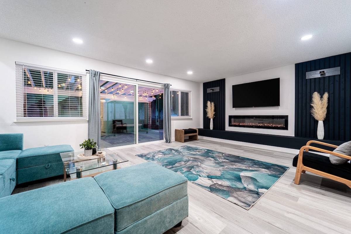A spacious living room features a soft turquoise sectional sofa, a glass coffee table, and an electric fireplace. Large windows provide natural light and a view of the outdoor area, while modern decor and a prominent area rug add visual interest to the space.