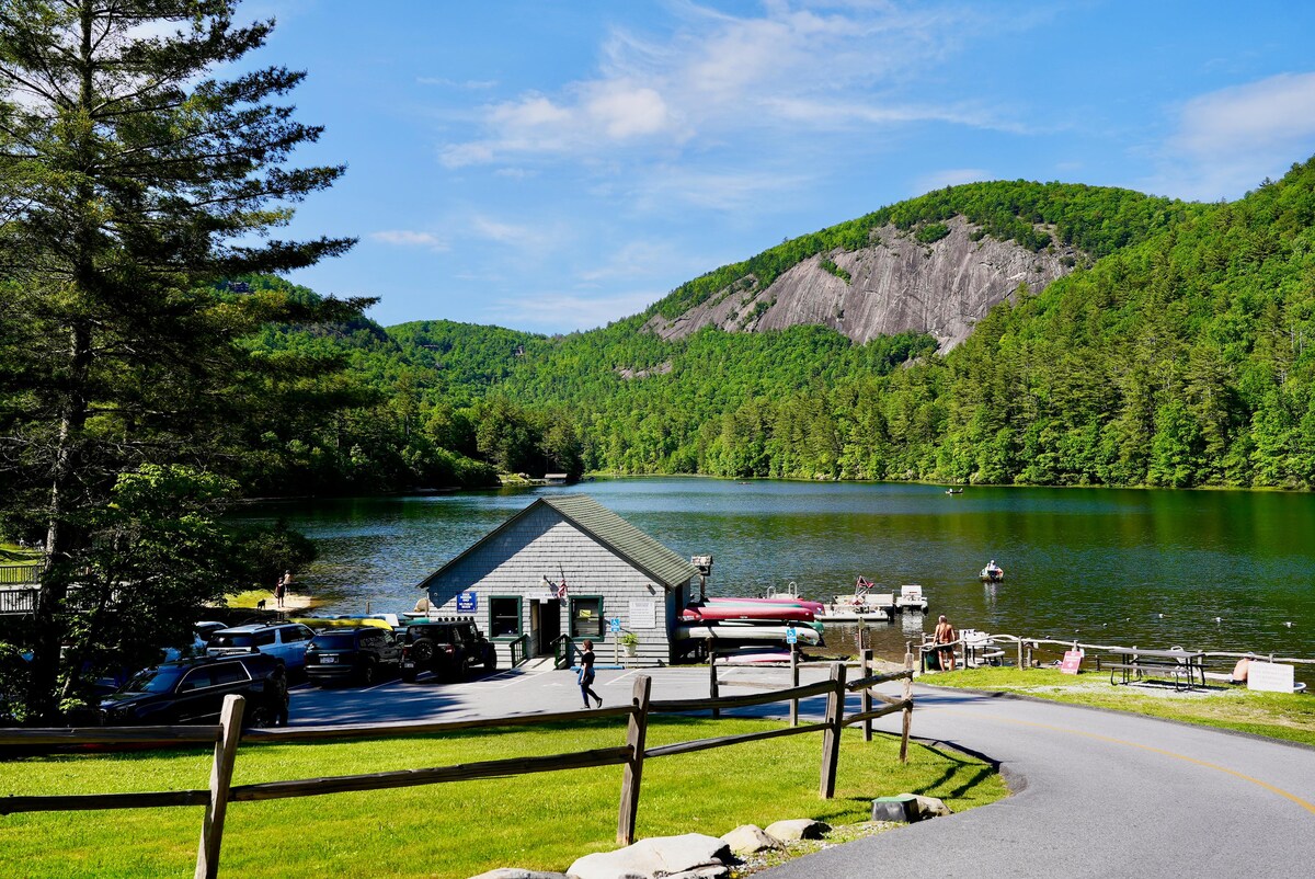 A serene lakeside scene is presented, featuring a rustic boathouse surrounded by lush greenery. Boats are docked at the water's edge, with rolling hills and a clear sky in the background. Gravel pathways and parked vehicles are visible along the well-maintained road.