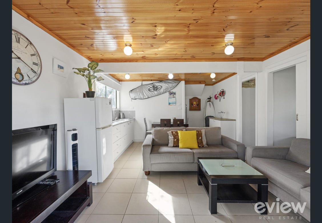 A bright and airy living area features a combination of comfortable seating, including a sofa with decorative cushions. A coffee table sits in front of the seating, and kitchen appliances are visible in the background. Natural light filters through the windows, enhancing the space.