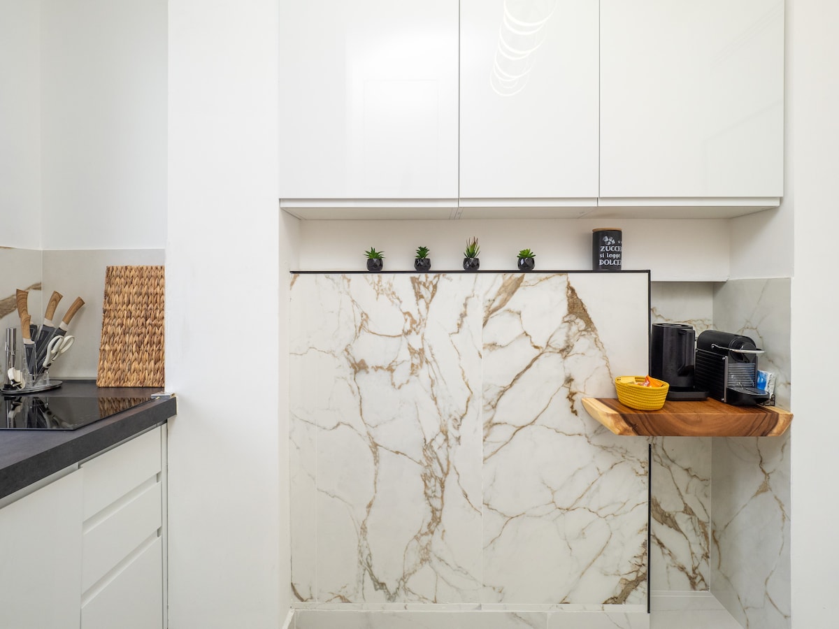 The kitchen features elegant marble backsplash complemented by sleek white cabinetry. A wooden shelf is mounted against the wall, displaying a small bowl, while kitchen utensils and appliances are neatly arranged on the counter, creating a functional cooking space.