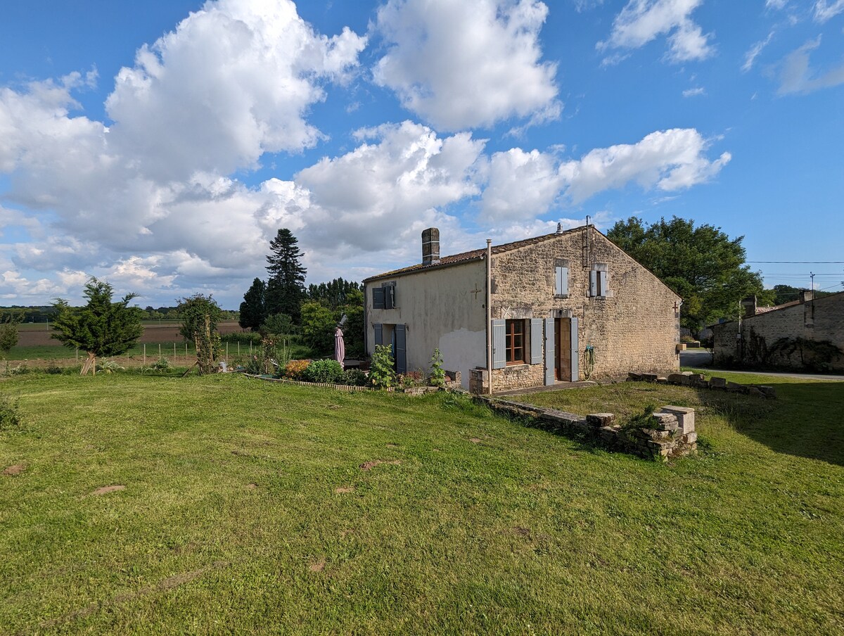 A charming stone home is set against a backdrop of rolling fields and blue skies. The structure features multiple windows and a rustic exterior, while a spacious grassy yard surrounds the property, dotted with shrubs and trees, providing a serene outdoor space.
