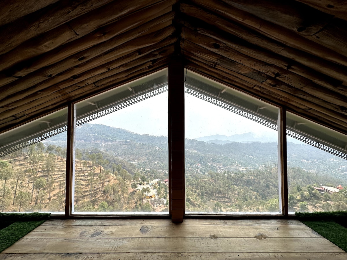 A spacious loft area features large triangular windows that frame a panoramic view of the surrounding Kasauli Hills. The natural wood ceiling complements the serene landscape, showcasing greenery and distant mountains in a tranquil setting.