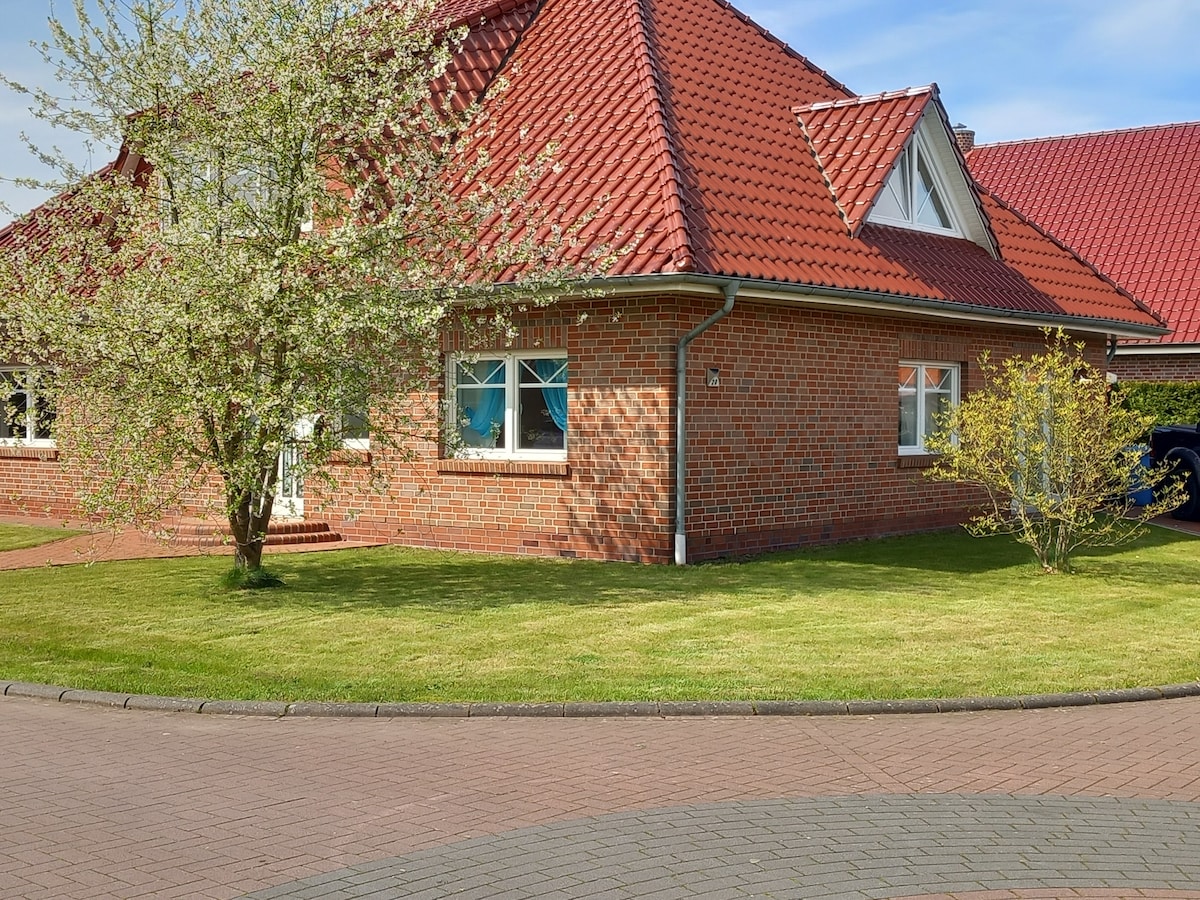 A charming brick house with a red tiled roof is surrounded by a well-maintained garden. A flowering tree and manicured grass enhance the outdoor space, while large windows allow natural light to fill the interior.