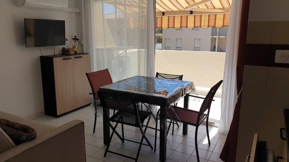 A dining area is presented with a table surrounded by four folding chairs. Light streams through sheer curtains, illuminating the space. A flat-screen television is mounted on the wall to the left, while a sideboard provides additional storage. An awning is visible outside, adding to the comfort.