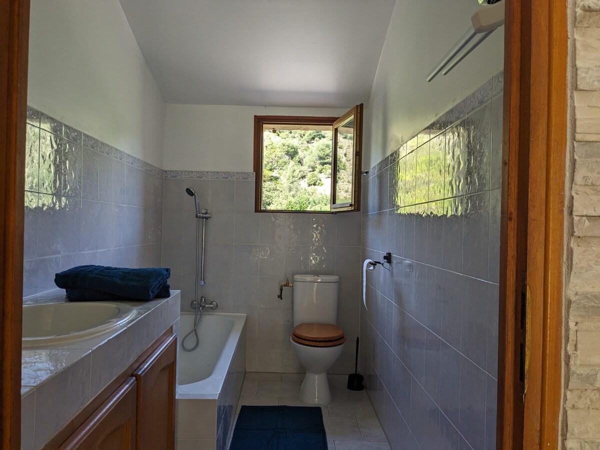 The bathroom is lined with light tiles and features a window allowing natural light to brighten the space. A bathtub is positioned beside the wall, while a toilet is located across from it. Fresh towels are neatly placed nearby.