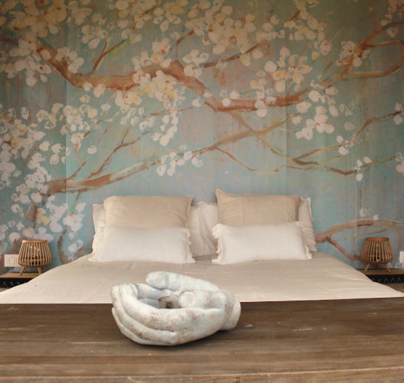 A serene sleeping area is highlighted by a textured mural of blooming branches against a soft blue background. The spacious bed features two cream-colored pillows and a white bedding set. A wooden table with a unique sculptural centerpiece completes the tranquil atmosphere.
