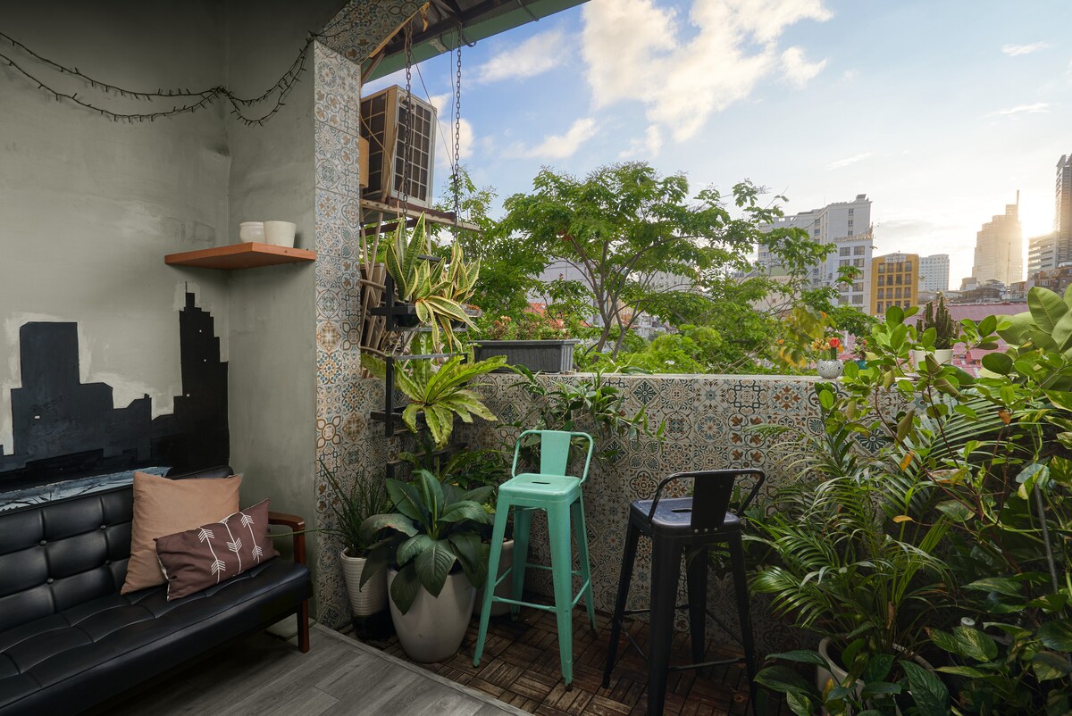 A cozy balcony area is furnished with a black chair and an aqua blue chair. Potted plants are placed around, providing greenery against a backdrop of city buildings. The sky is partly cloudy, with soft sunlight illuminating the space and revealing colorful tile patterns on the walls.