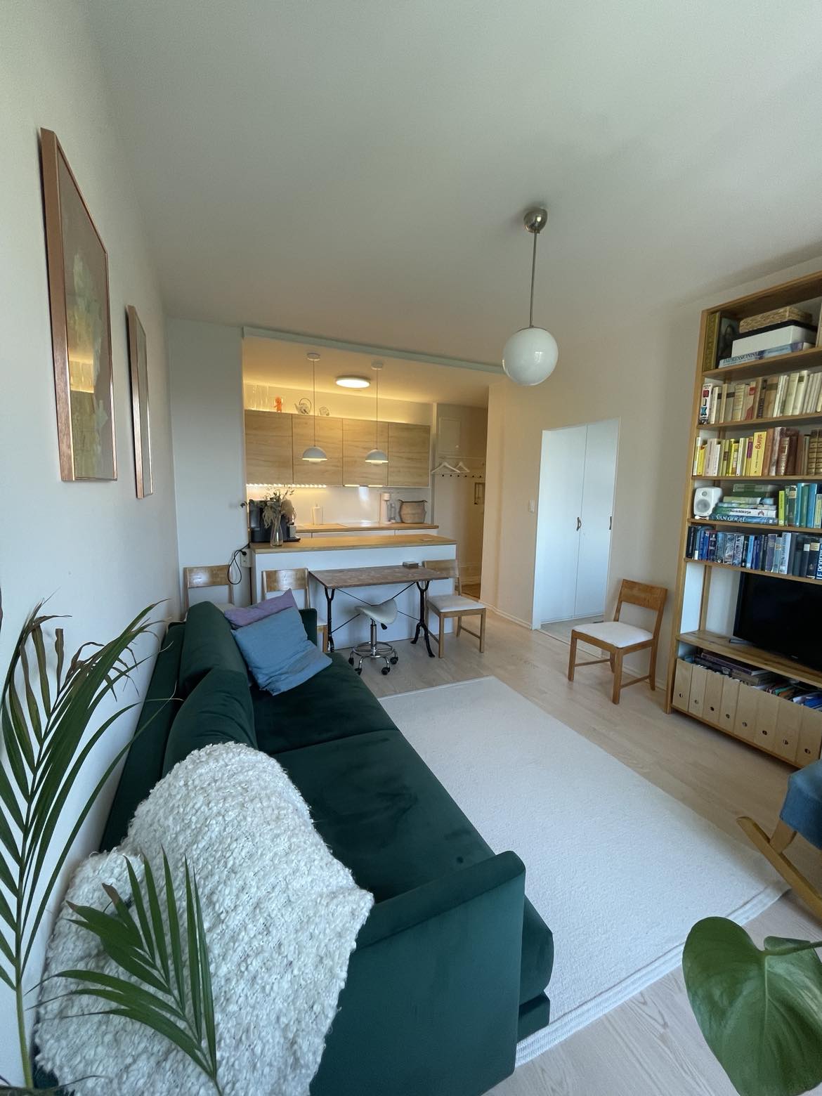 An open concept living area is presented, featuring a dark green sofa adorned with a light throw. A wooden dining table is visible along with four chairs. The kitchen is illuminated with pendant lights, while bookshelves line one wall, displaying various books and plants.