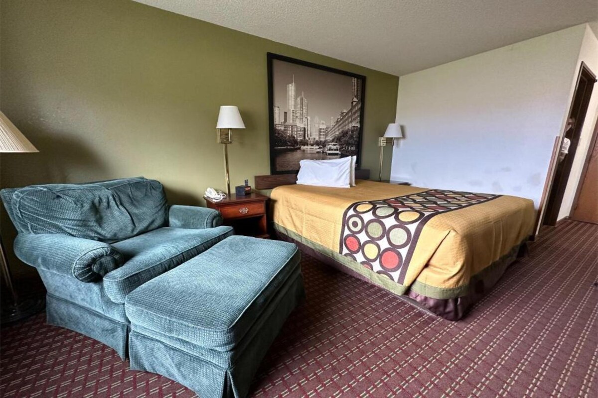 A clean accommodation is presented with a spacious bed covered in a colorful patterned blanket. A cozy upholstered chair and footrest are positioned nearby, accompanied by a small nightstand featuring a lamp and decorative items. A large framed photograph adds visual interest to the wall.