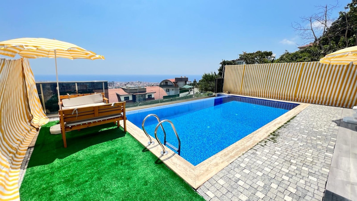 A private pool area features a well-maintained swimming pool with clear blue water, surrounded by light yellow and white striped sunshades. A wooden bench is positioned nearby on green grass, with a scenic view of the ocean and distant city visible beyond the pool.