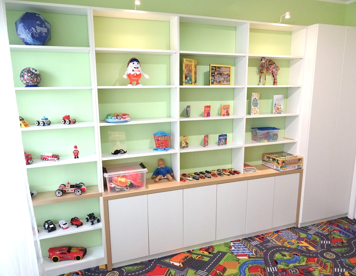 A brightly colored shelving unit is filled with various toys and books. Items include small vehicles, a plush toy, and games, arranged on white shelves against a light green wall. A vibrant, patterned carpet depicting a road design is visible below.