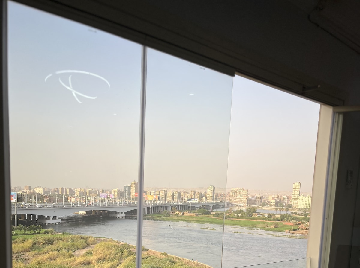 A large glass window offers a clear view of the river and city skyline. The landscape features a blend of greenery along the riverbank, with urban buildings visible in the distance. Soft natural light fills the interior space through the window.