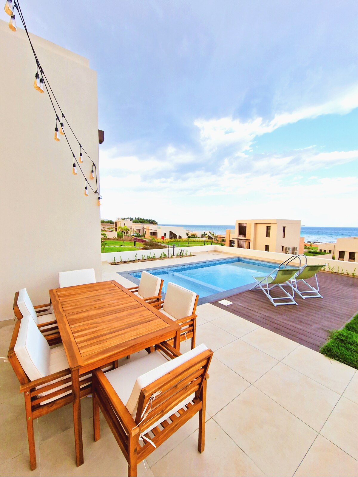 An outdoor dining area features a wooden table surrounded by multiple chairs. Soft string lights hang above, creating a relaxing ambiance. A private pool is visible beyond the patio, with views of the Arabian Sea and landscaped grounds enhancing the outdoor space.