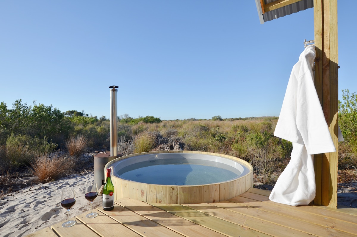 A wood-fired hot tub sits on a wooden deck, surrounded by natural vegetation. Nearby, a bottle of sparkling wine and two glasses are placed on the surface. A white bathrobe hangs casually on a post, with clear blue skies overhead.