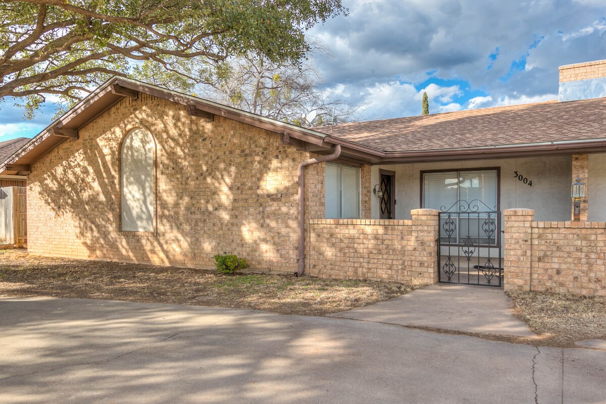The exterior showcases a single-story brick house framed by a large tree and an expansive sky. A decorative wrought iron gate leads to the entrance, while the well-maintained yard features a gravel pathway extending towards the door.