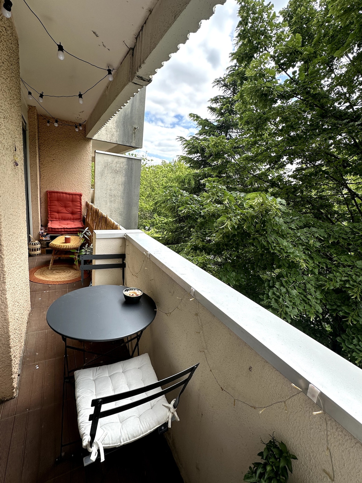 A cozy balcony offers a small round table accompanied by two chairs, one adorned with a bright cushion. Green foliage provides a natural backdrop, enhancing the serenity of the space. Soft lighting is strung along the railing, creating an inviting atmosphere.