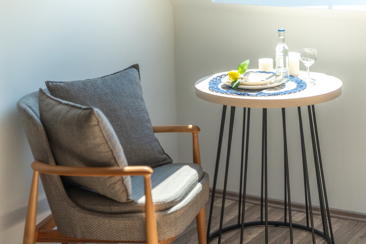 A comfortable chair is positioned next to a small round table. The table is adorned with a blue cloth and set with a plate, glassware, and a bottle. Natural light is streaming in from a nearby window, creating a bright atmosphere.