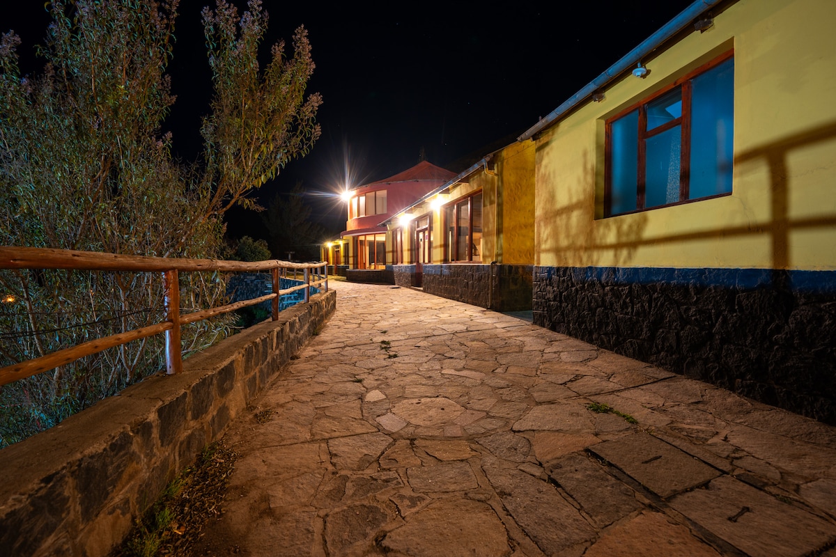 A stone pathway is illuminated by warm lights, leading to a vibrant yellow building. Wooden railings accompany the walkway, while trees are visible along the edges. The setting conveys a sense of calmness against the night sky.
