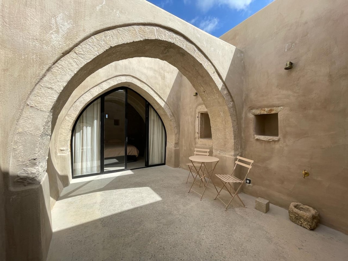 An outdoor patio area is framed by stone arches and features a small table with two chairs. Large glass doors lead into the living space, allowing natural light to illuminate the textured walls. A peaceful atmosphere is created by the simplicity of the design.